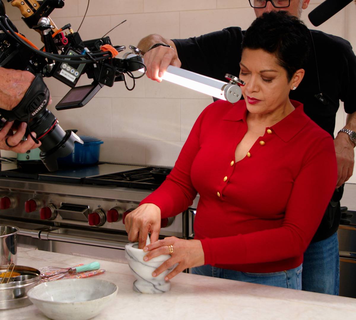 Anupy Singla in her Chicago kitchen wearing a red shirt and grinding spices with a mortar and pestal. She is surrounded by a film crew with lights and cameras.